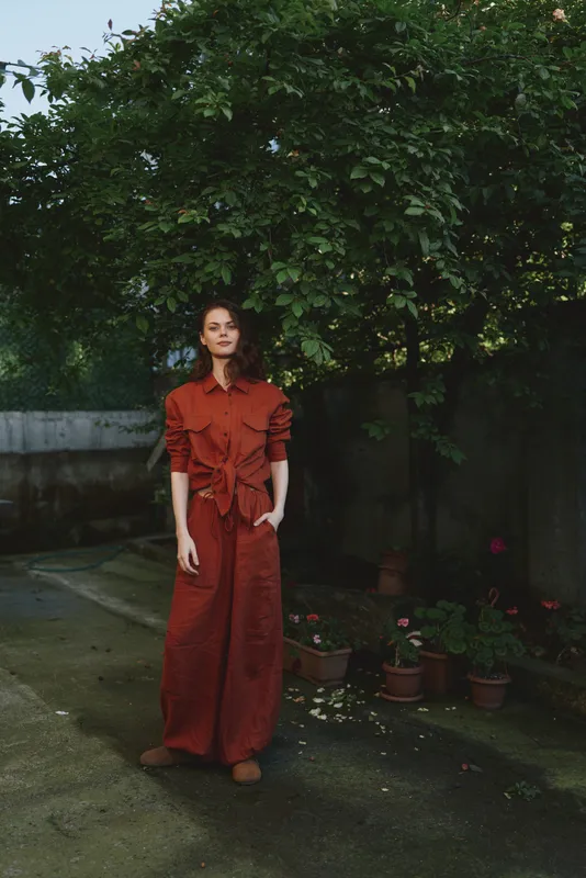 Woman in a red jumpsuit stands in a lush garden, illustrating contemporary outdoor lifestyle and fashion in a natural setting, with soft light and calm mood for editorial use
