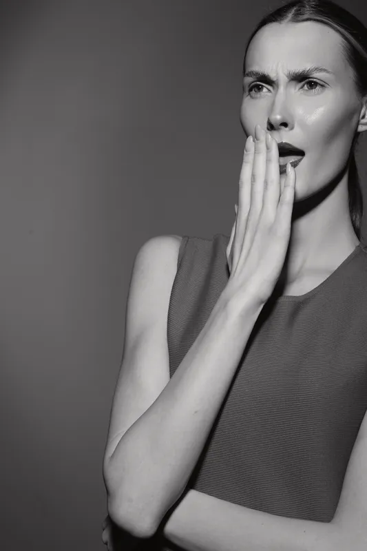 Young Caucasian woman with light skin looks surprised and covers mouth with hand in studio lighting.