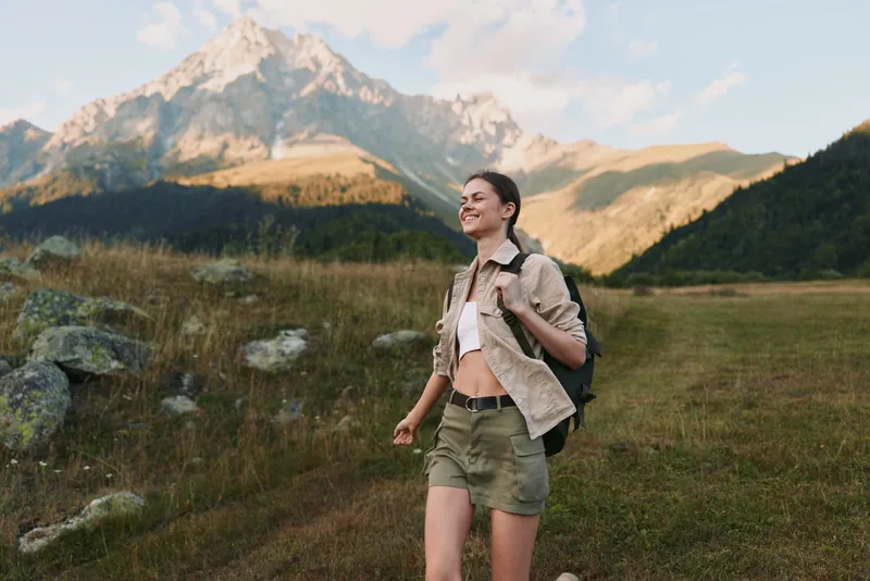 A woman hiker walks through a bright meadow toward distant mountains, wearing shorts, a light shirt, and a backpack. Concept: outdoor exploration and active lifestyle.