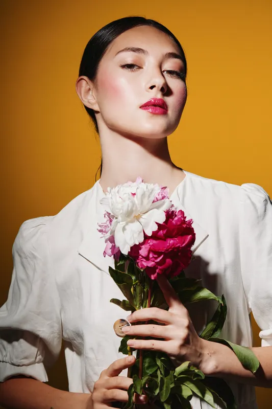 Young fashionable woman with flawless makeup and red lips holds fresh flowers against a bright yellow background. Beauty portrait emphasizes elegance and creative fashion concept.