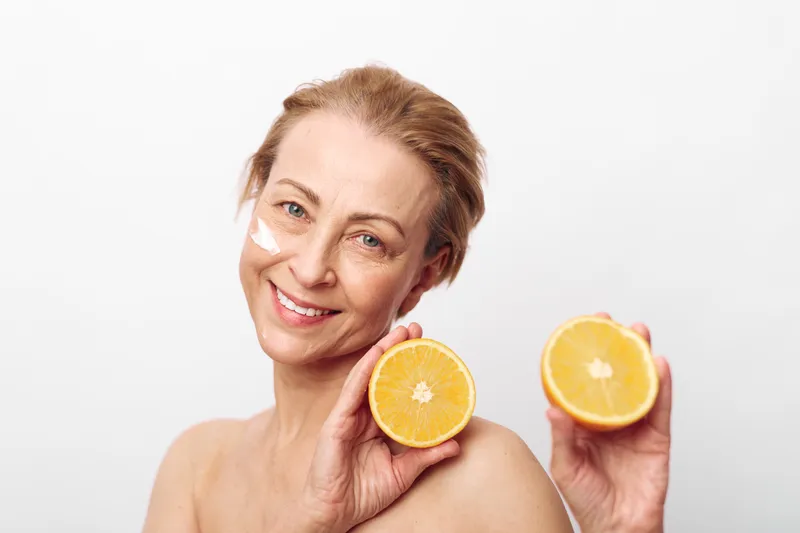Smiling mature woman with facial cream on cheeks holds two halves of fresh orange, isolated on white background. Concept of natural skincare, healthy lifestyle, and citrus beauty treatment.