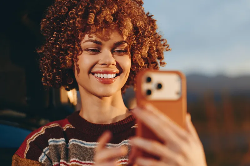 Happy woman lifestyle selfie outdoors at sunset, curly red hair and bright smile, holding a phone toward mirror camera, casual knit sweater, warm evening light, candid street moment.