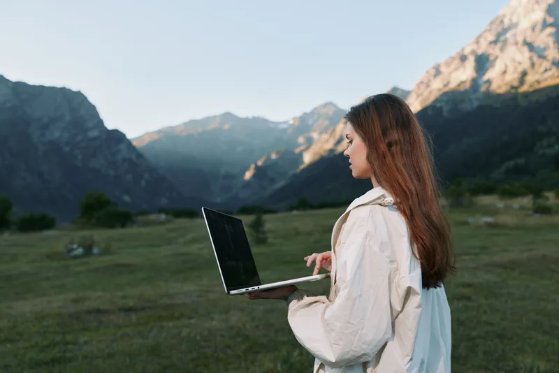 A focused young woman stands in a scenic valley, holding a laptop as she works remotely. She looks attentive and calm, blending nature with modern digital tasks.