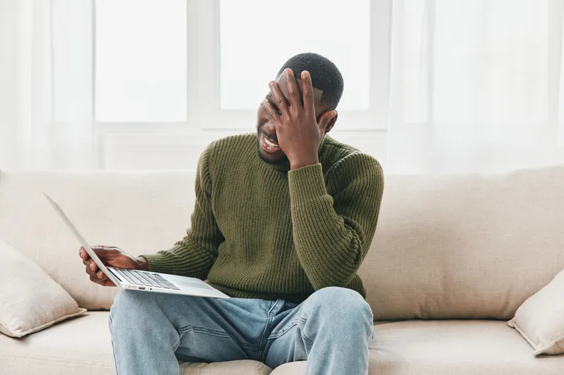 Young Black man expressing frustration while working on a laptop at home, wearing a cozy green sweater, in a bright and airy living room environment