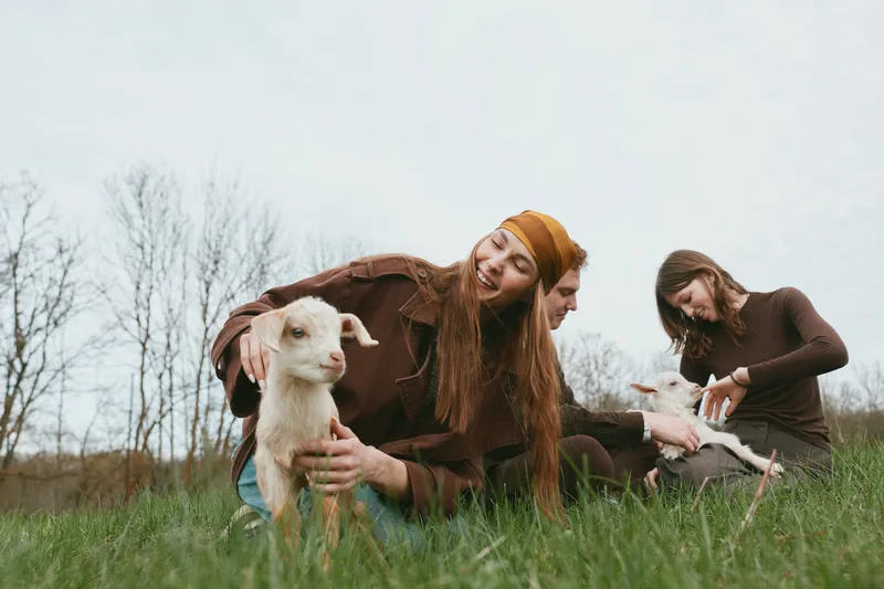 Lifestyle scene with three friends in a rural field, interacting with a playful goat kid, outdoor mood, nature, countryside vibe, and a cozy, candid moment of care and curiosity
