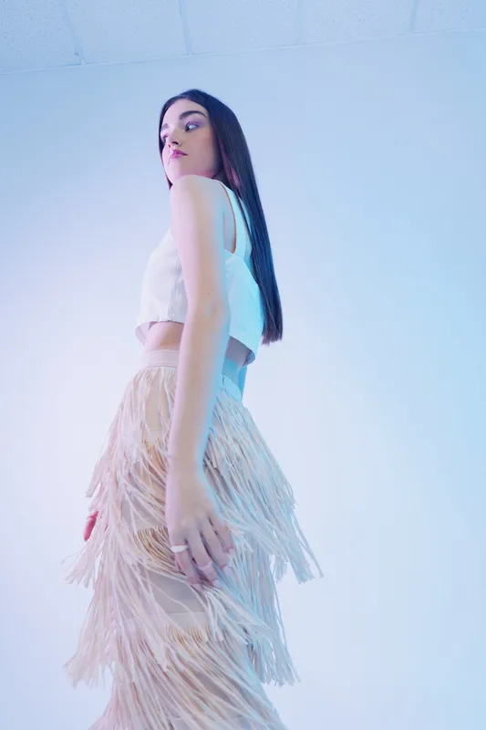Young woman beauty fashion model poses in a white crop top and fringed skirt against a light background. Concept for style, elegance, and modern clothing trends.