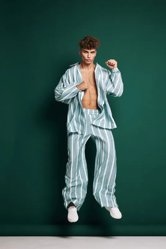 Young man jumping in striped pajamas with open shirt and white sneakers on green background. Casual fashion, energetic pose and modern style for lifestyle and comfort.