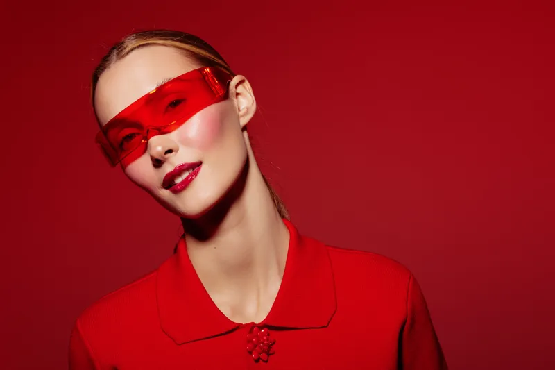 Young woman with red sunglasses and red outfit poses against red background. Fashion portrait with glossy lips and sleek hairstyle. Concept of style, color, and modern beauty.