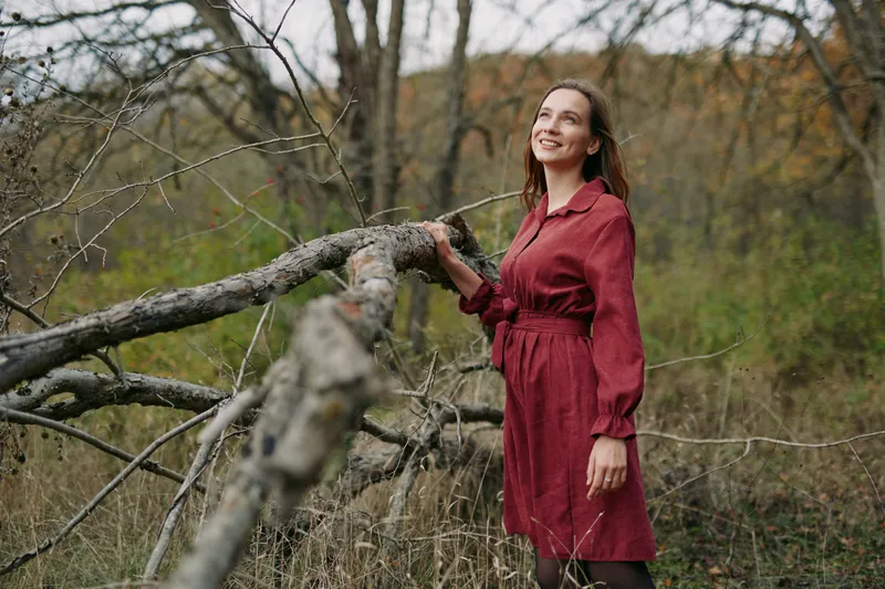Woman in a simple red dress stands by a fallen tree in a natural meadow, smiling and looking up, conveying grounded authenticity and quiet credibility in a candid outdoor portrait.