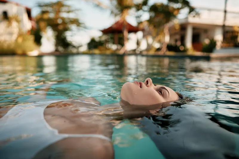 Young woman relaxing in a swimming pool with eyes closed. Lifestyle concept of leisure and wellness in a sunny outdoor setting. Calm water reflects a peaceful mood.