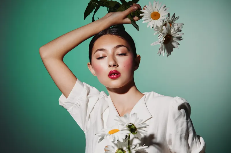 Young woman in white blouse with bright makeup poses creatively holding large white flowers against soft turquoise background. Concept for beauty, fashion, and floral art.