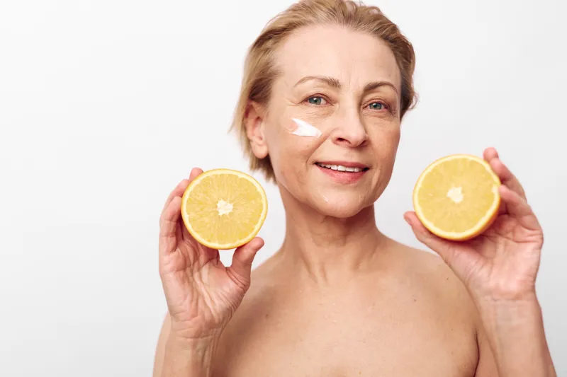 Mature woman with short blonde hair holds two halves of orange and applies facial cream on cheek. Concept for natural skincare, healthy lifestyle, and aging beauty care at home.