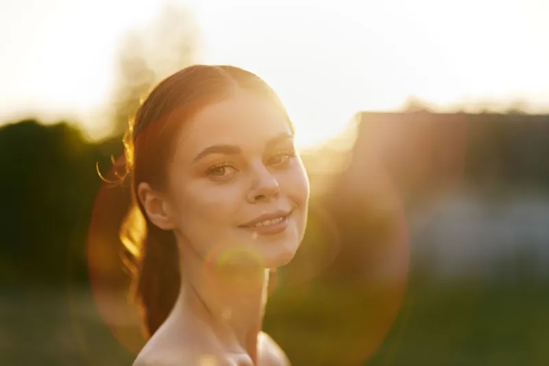 Young woman smiling softly in a sunlit outdoor setting during golden hour, capturing the essence of joy and natural beauty