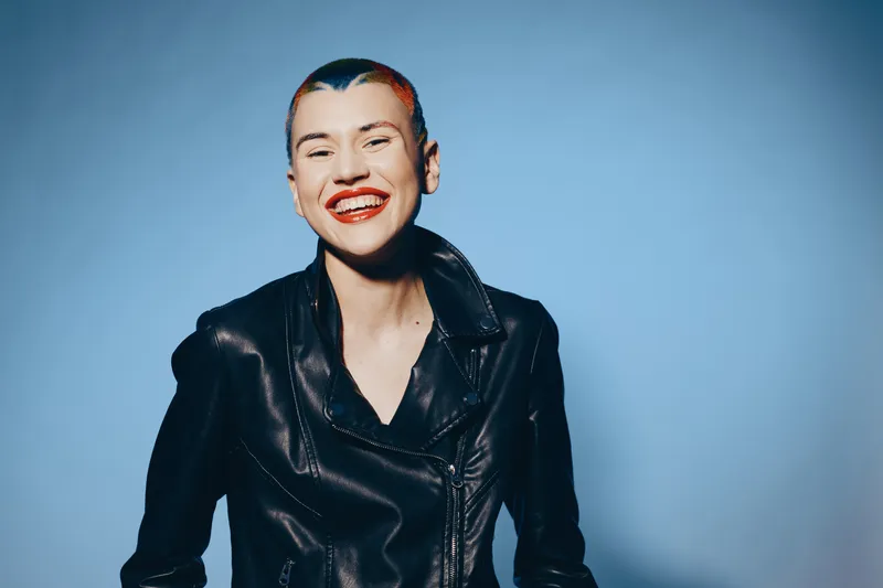smiling woman with short hair wearing black leather jacket and bright red lipstick, joyful expression, casual style, blue background, studio lighting