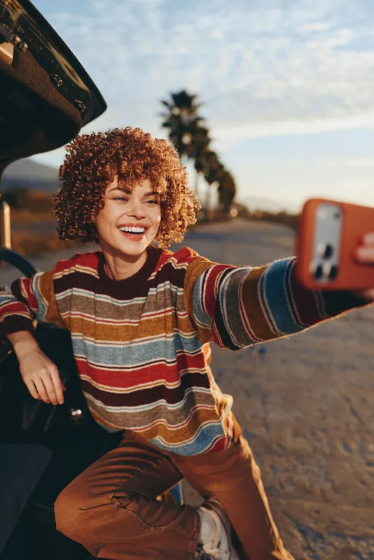 Woman with curly hair smiling outdoors taking selfie with phone by car on coastal street, summer lifestyle, warm sunlight, relaxed travel moment, candid portrait, joyful expression, carefree lifestyle