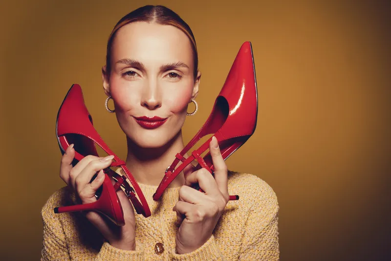 Smiling woman holds red high heel shoes near face on yellow background. Fashion concept with stylish footwear and confident female model in warm lighting studio portrait.