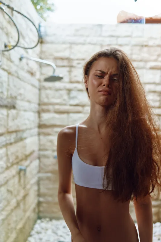 Young woman with long curly hair wearing white sports bra feeling uncomfortable in outdoor shower with stone wall background on sunny day.