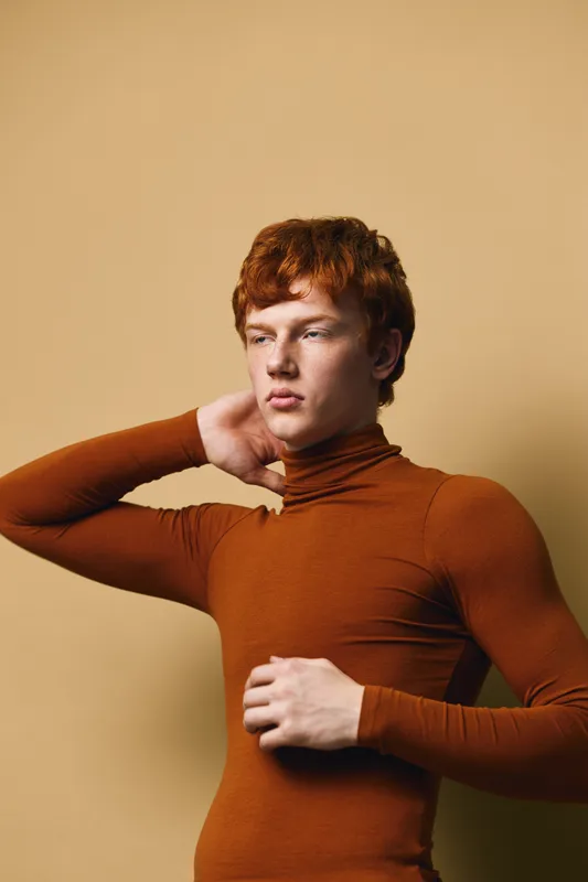 Young man with red hair posing in brown turtleneck sweater against beige background with thoughtful gaze and hand touching neck in studio portrait.
