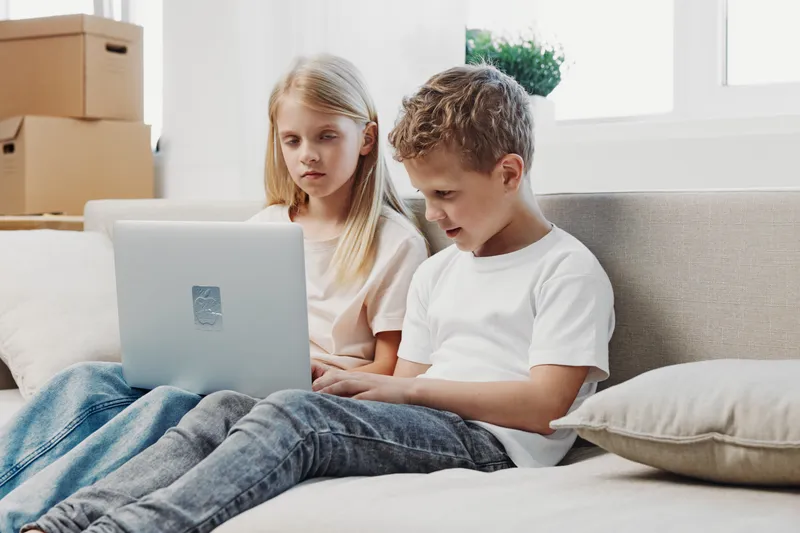 Two children sitting on sofa using laptop together, focused on screen in bright living room. Concept of learning, technology, communication, and childhood development.