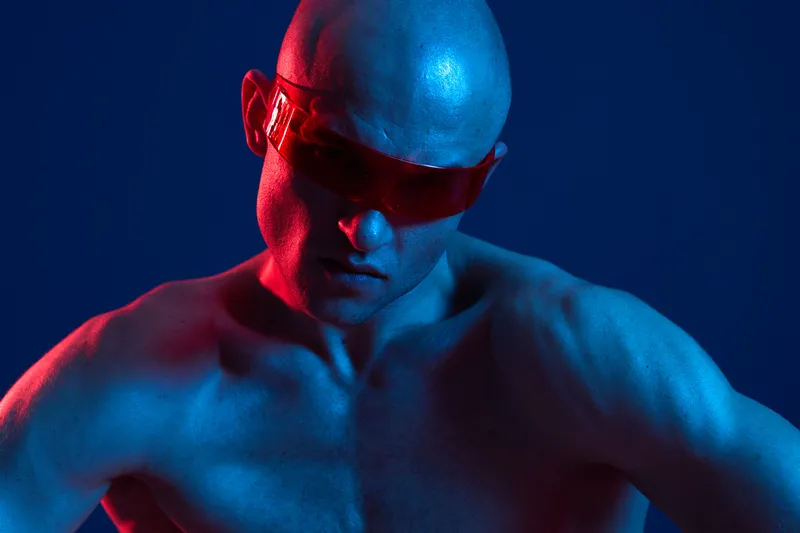 A focused man in red sunglasses poses dramatically with a blue and pink background, showcasing his athletic physique and intense expression. Black and white concept.