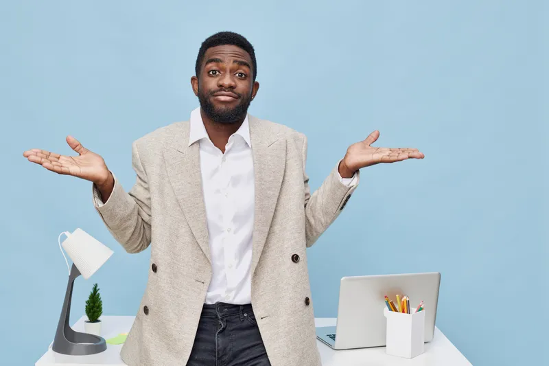 Confused man with raised hands standing at modern office desk on plain blue background. Professional workplace scenario, uncertain expression, business attire, workspace setup, confident, casual style