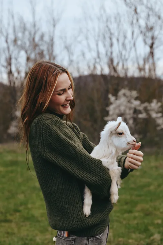 A woman in a dark green sweater holds a young goat kid in a sunny open field, smiling softly. A warm, rustic moment capturing rural lifestyle and gentle animal bonding.