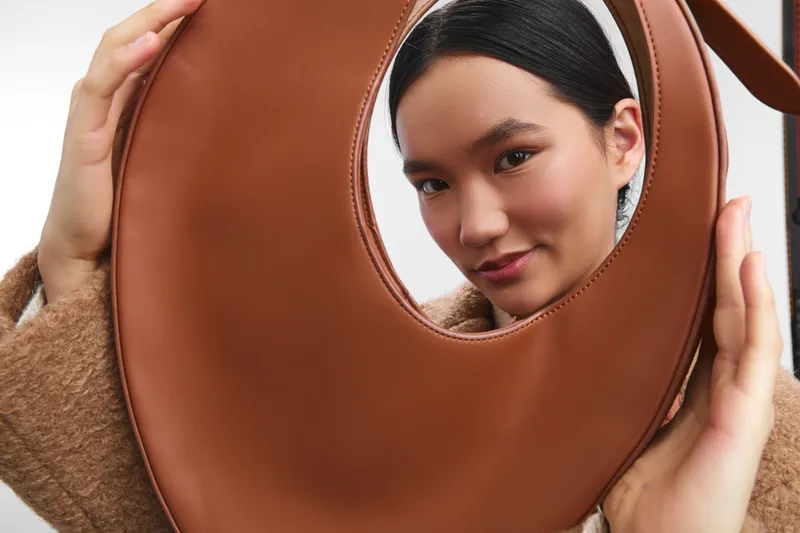 Asian woman peers through a round fashion bag, posing in a softly lit studio. She smiles with curious charm, creating a chic, playful and inviting visual mood.
