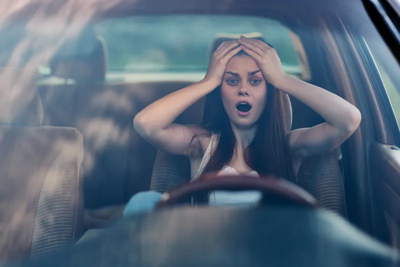 A shocked young woman sitting in her car with her hands on her head, depicting a moment of anxiety and surprise against a blurred outdoor background.