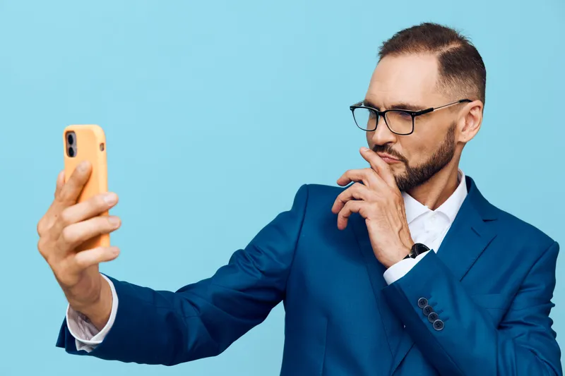 A professional businessman poses against a solid blue background, checking a smartphone with thoughtful focus. He wears a sharp suit, conveying confidence and curiosity, signaling leadership,
