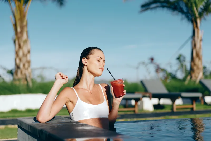 Relaxed poolside scene shows a woman in a white crop top sipping a red drink through a straw, enjoying outdoor sunshine, casual leisure and summer lifestyle by the water.