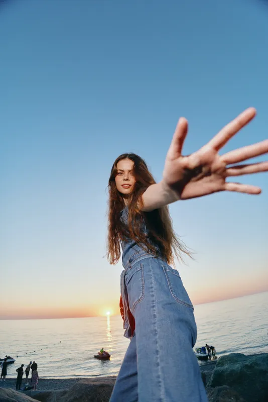 young woman reaching hand towards camera at seaside during sunset with clear blue sky and relaxed mood