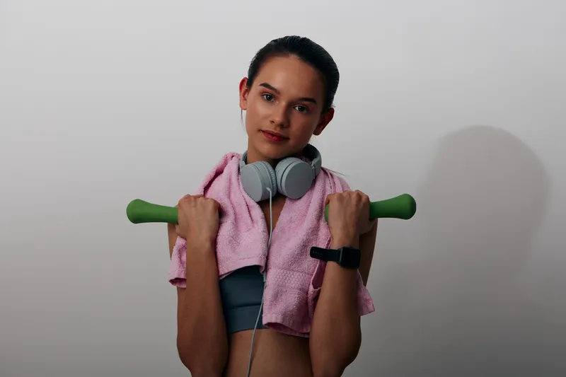 Woman fitness in a gym setting, holding green dumbbells with a pink towel around shoulders and headphones, showcasing workout energy, training discipline and confident sport mindset.