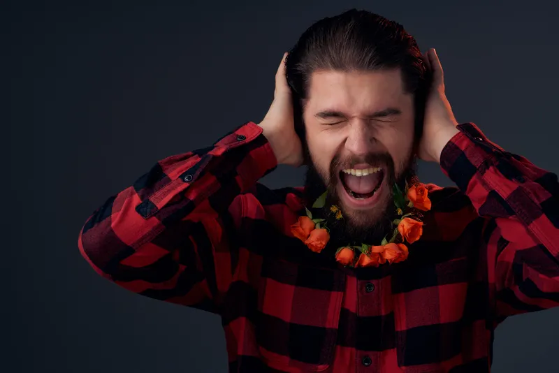 Бородатый мужчина с цветами, вплетёнными в бороду, одет в клетчатую рубашку на тёмном фоне