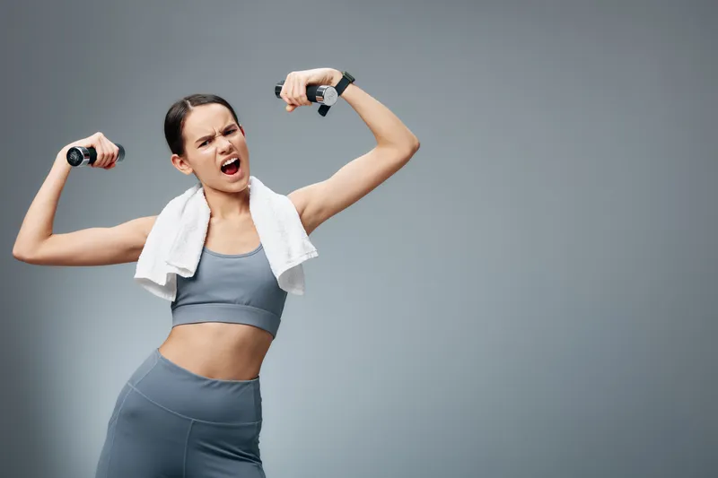 Fitness woman in athletic outfit lifts light dumbbells with a towel over her shoulders, standing in a clean studio. Dynamic pose conveys strength, cardio focus, and an active, healthy lifestyle.
