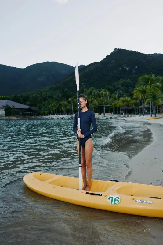 Young woman in a wetsuit stands barefoot on a bright yellow kayak at the water's edge, calm sea beside a sandy shore and lush hills, evoking adventure and summer serenity.