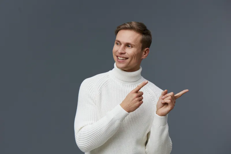 Cheerful man in white sweater pointing sideways studio portrait on gray background smiling relaxed casual fashion concept