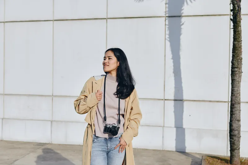 Woman with camera in a beige coat and pink sweater stands outdoors beside a light wall, showcasing street style, fashion, travel vibe, and candid urban lifestyle