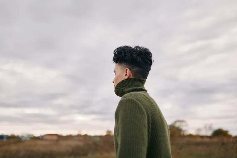 A young man in a green turtleneck stands in a windswept field, profile against an overcast sky, conveying authenticity and credibility through a natural, unposed portrait with calm, contemplative