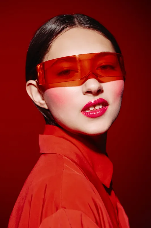 Fashion portrait of a woman wearing transparent red sunglasses and glossy red lipstick. Her makeup features bold blush, set against a saturated red background, highlighting beauty and creativity.