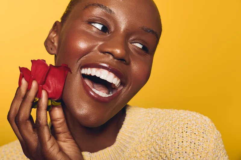 Happy African woman with dark skin laughing and holding red rose near face on yellow background. Concept of joy, beauty, skincare, and positive emotions in studio portrait.