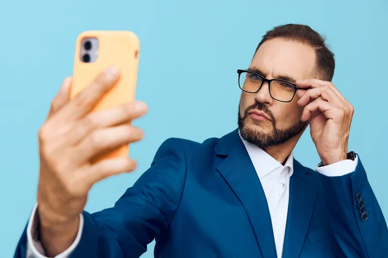 A business professional man poses for a selfie against an isolated blue background, exuding confidence and focus while adjusting his glasses and checking his phone.
