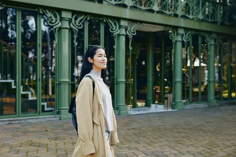 Woman in a beige coat walking along a city street with a backpack, natural light, candid style, casual fashion, urban ambiance, storefront windows, calm mood, everyday travel scene.