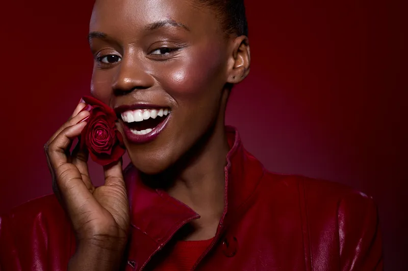 Smiling black woman with dark skin holds red rose near face wearing red leather jacket. Close-up portrait on red background. Concept of beauty, joy, fashion, and natural skin glow.