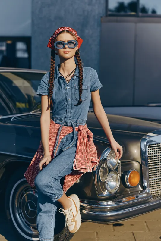 Lifestyle woman in modern boho-western denim street style leaning on vintage car with film color effect and casual accessories under bright sunlight.