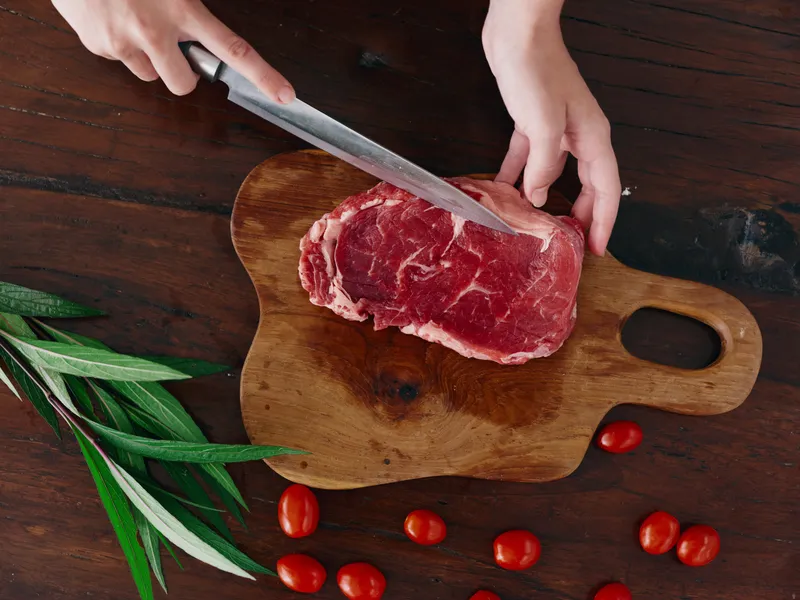 Женщина с ножом в руке нарезает свежее мясо стейка для жарки на кухне; на столе соль, перец и другие специи, красные помидоры черри и зелень; приготовление ужина в домашней кухне