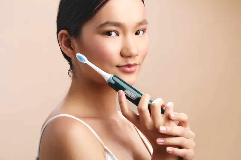 asian woman smiles softly while brushing teeth with a modern electric toothbrush in a clean bathroom, presenting a calm, healthy routine and inviting personal care moment.