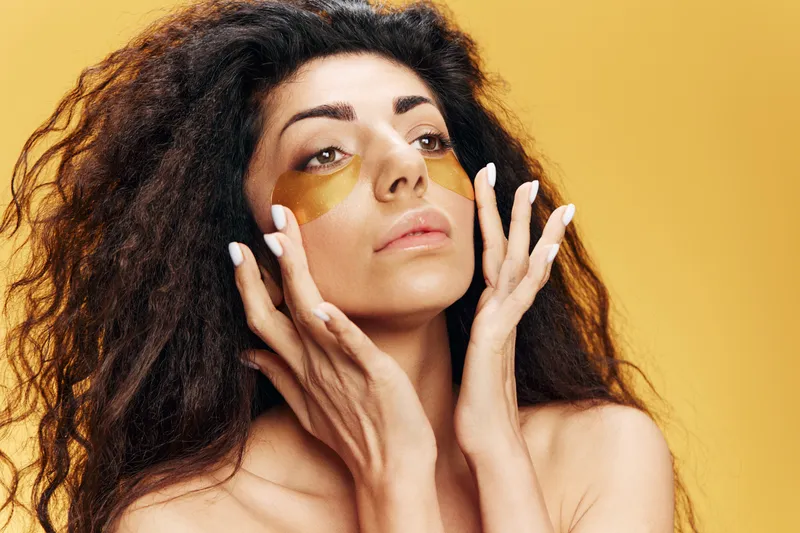 Красивая симпатичная латиноамериканка с золотыми гидрогелевыми патчами под глазами увлажняет кожу лица, смотрит в сторону, позирует изолированно на желтом фоне
