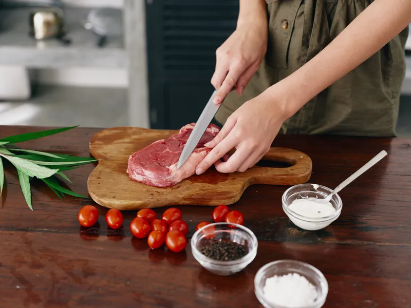 Женщина с ножом в руке нарезает мясо стейка для жарки на кухне