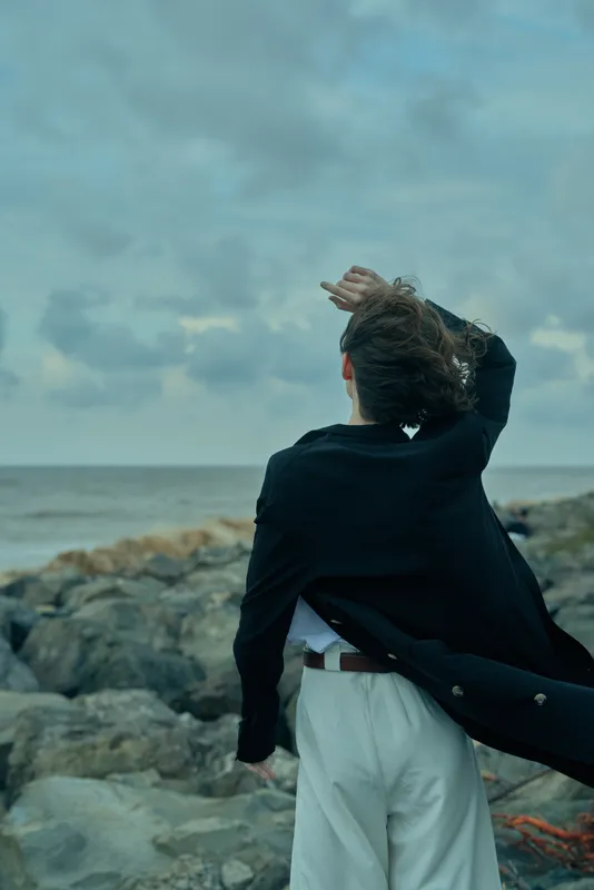 Wind swept coastline scene with a silhouette facing the sea, coat fluttering, rocky shore, open horizon, moody light, outdoor adventure, solitude, exploration, sea breeze, travel vibe