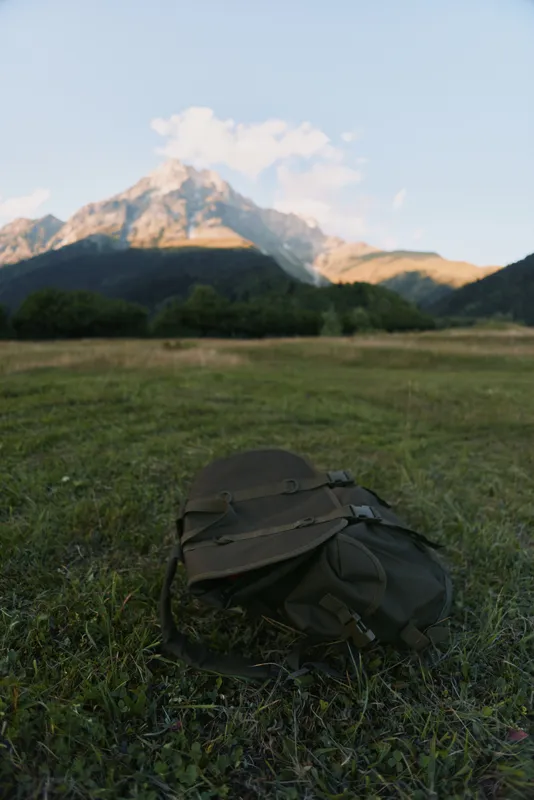 A lone backpack sits on a grassy meadow with towering mountains in the background, capturing a moment of outdoor exploration and quiet solitude.
